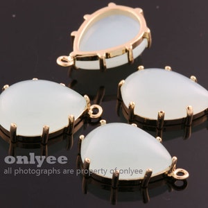 Könnte beinhalten: Vier weiße tropfenförmige Glascabochons mit goldenen Metallfassungen. Jeder Cabochon hat eine kleine Schlaufe oben zum Anbringen an Schmuck.