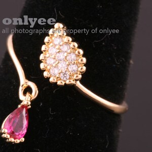Puede incluir: Un anillo de oro con un diseño en forma de lágrima adornado con un grupo de piedras de circonita cúbica transparentes. Una piedra roja en forma de lágrima colgante cuelga del anillo.