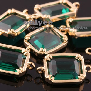 May include: A group of emerald green glass rectangular charms with gold-toned metal frames and loops. The charms are scattered on a black surface.