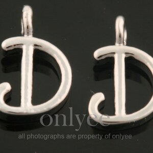 Könnte beinhalten: Zwei silberne Metall-Anhänger in Form des Buchstabens D mit einer Öse oben zum Aufhängen. Der Buchstabe D ist in einer kursivschrift.
