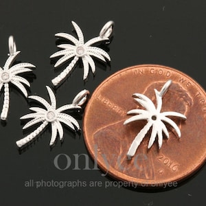 May include: Four silver palm tree charms with clear rhinestones. The charms are shown next to a US penny for size comparison.  "In God We Trust" and "Liberty" are visible on the penny.