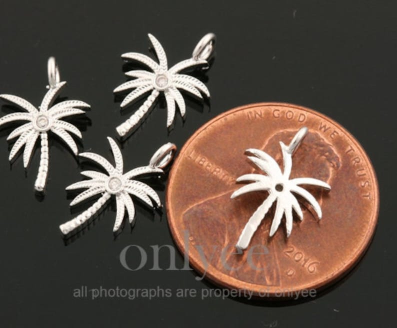 May include: Four silver palm tree charms with clear rhinestones. The charms are shown next to a US penny for size comparison.  "In God We Trust" and "Liberty" are visible on the penny.