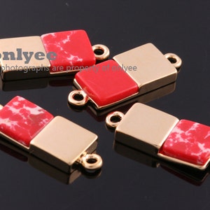 Pode incluir: Achados de joalharia retangulares dourados com uma pedra quadrada vermelha e branca salpicada. Cada achado tem uma argola em cada extremidade para fixar a outros componentes. A imagem mostra vários destes achados.