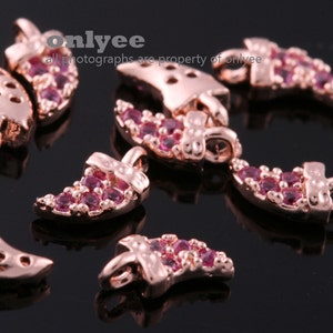 May include: A group of rose gold-coloured metal charms shaped like a crescent moon with pink rhinestones. The charms have a loop at the top for attaching to jewellery.