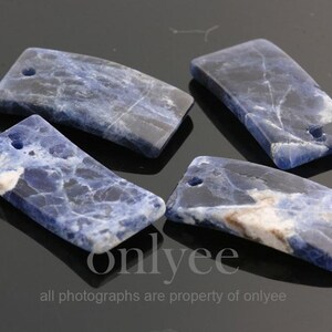 May include: Four blue and white rectangular pieces of polished sodalite stone with holes drilled through them. The stones are arranged on a black surface.