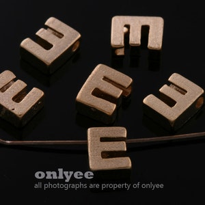 Puede incluir: Primer plano de siete abalorios de metal de color dorado en forma de letra E. Los abalorios están en varias posiciones sobre una superficie negra. Los abalorios son todos del mismo tamaño y forma, y tienen una superficie ligeramente texturizada.