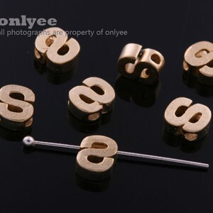 May include: Seven gold-colored metal letter "S" beads with a hole through the center of each bead. The beads are arranged on a black surface with a silver metal rod.