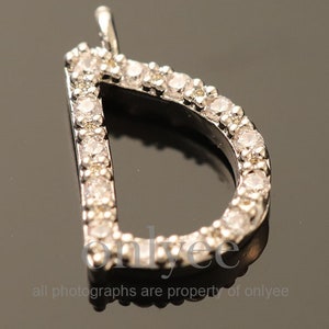 Könnte beinhalten: Ein silberner Anhänger in Form des Buchstabens D, mit kleinen klaren Zirkonia-Steinen, die um den Rand herum gesetzt sind.