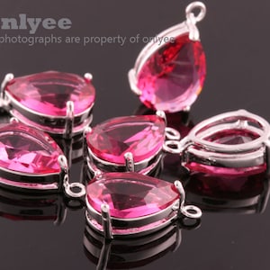 Pode incluir: Seis pingentes de cristal rosa em forma de lágrima com engastes prateados e argolas de salto. Os pingentes estão dispostos sobre uma superfície preta.