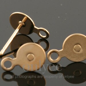 May include: Two gold-toned earring backs with a small loop on the side. The earring backs are round and have a flat surface.