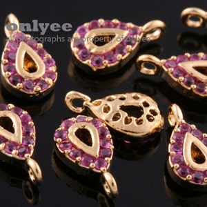 May include: A group of eight gold-tone teardrop-shaped charms with pink rhinestone accents. Each charm has a loop at the top for attaching to a necklace, bracelet, or other jewelry.