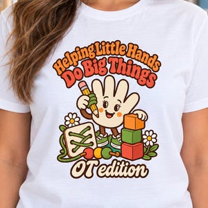 May include: A white short-sleeve t-shirt featuring a colorful graphic of a cartoon hand holding a pencil next to building blocks and a lacing toy. The text reads Helping Little Hands Do Big Things OT edition in a retro orange and brown font.