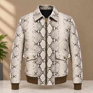 May include: A bomber jacket featuring a cream and dark gray snakeskin pattern. The jacket has a pointed collar, a central brass-toned zipper, two front flap pockets with brass-toned buttons, and dark brown ribbed cuffs and hem.