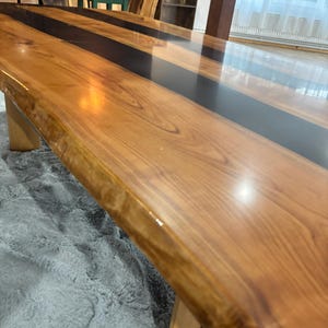 Puede incluir: Una mesa de comedor de borde natural con una superficie de madera pulida y una incrustación central prominente de resina negra lisa y opaca. La mesa tiene un acabado brillante que refleja la luz, resaltando la veta de la madera y el divisor negro.