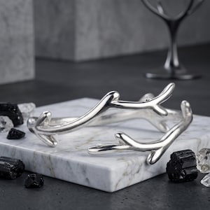 May include: A silver-toned cuff bracelet shaped like two intersecting tree branches with smooth, polished surfaces. The bracelet features a bypass design with open ends, showcasing a metallic finish that reflects light across its curved, organic form.