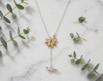 May include: A silver chain necklace featuring a gold-toned sun charm and a small silver figure pendant dangling from the center. The necklace is designed as a lariat style piece with a delicate link chain and a polished metallic finish.