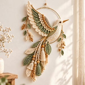 Puede incluir: Un adorno de pared con un pájaro tejido a ganchillo en color crema, verde salvia y amarillo mostaza, montado sobre un aro de metal dorado. El pájaro está decorado con hojas tejidas, cuentas de madera y borlas colgantes a juego.