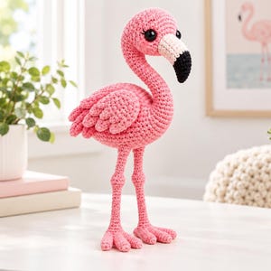 May include: A crocheted flamingo figurine crafted from bright pink yarn. The bird features a long, curved neck, textured wing details, and a beak colored in sections of white and black. It has large, round, black plastic eyes and two long legs.