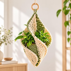 Puede incluir: Un soporte colgante para plantas hecho de hilo de ganchillo en secciones alternas de color crema y verde oliva. La estructura presenta un diseño en espiral que sostiene una pequeña maceta verde con una planta de hojas verdes, suspendida por un aro de madera.