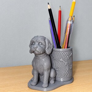 Op de afbeelding: Een grijze 3D-geprinte bureau-organizer met een gebeeldhouwde hond die naast een cilindrische pennenhouder zit. De pennenhouder is versierd met een golvend patroon en kleine botvormen. Verschillende gekleurde potloden en pennen zitten erin.