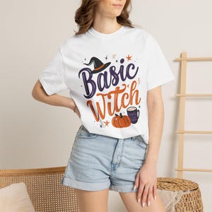 May include: A white short-sleeved t-shirt featuring a graphic print with the text Basic Witch in purple and orange script. The design includes a black witch hat, an orange pumpkin, and a dark mug with text, accented by small stars.