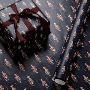 May include: A dark navy blue wrapping paper featuring a repeating pattern of small, colorful nutcracker figures arranged in vertical rows, accented by thin gold lines. A square gift box wrapped in the same paper is tied with a dark maroon ribbon.