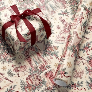 May include: A gift box wrapped in cream-colored paper with a red ribbon, alongside a matching roll of wrapping paper. The paper features a detailed red and green illustration of nutcrackers, dancers, harps, and holiday greenery on a cream background.