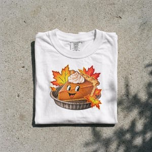 Op de afbeelding: Een wit t-shirt met een afbeelding van een cartoon pompoentaartpunt met een lachend gezicht en een toefje slagroom, omringd door oranje en gele herfstbladeren van een esdoorn. Het shirt heeft een Comfort Colors merklabel in de binnenhals.