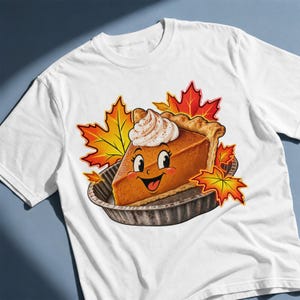 以下が含まれることがあります： 白いTシャツに、ホイップクリームをのせて笑顔を浮かべたパンプキンパイのイラストが描かれています。パイは金属製の型に入っており、鮮やかなオレンジと黄色の秋の紅葉したカエデの葉に囲まれています。