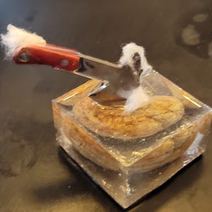 May include: A clear, square resin block containing a tan, textured stone. A miniature metal hatchet with a red handle is embedded into the top of the resin, with small tufts of white cotton-like material attached to the handle and the blade edge.