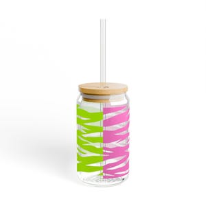 Puede incluir: Un vaso de vidrio transparente de forma cilíndrica con un patrón de rayas de cebra dividido en colores verde lima brillante y rosa intenso. El vaso incluye una tapa de bambú de color claro con un orificio central y una pajita de plástico transparente.