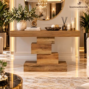 May include: A rectangular wooden console table with a thick top and a base composed of three stacked, offset wooden blocks. The table surface holds a speckled vase with white flowers, a gold nautilus shell, two books, a bowl, and candles.
