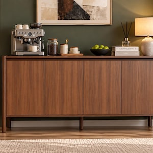 May include: A rectangular wooden sideboard featuring four vertical cabinet doors with a smooth walnut-toned finish and rounded side edges, supported by cylindrical legs, providing a flat surface for a coffee machine and decorative items.