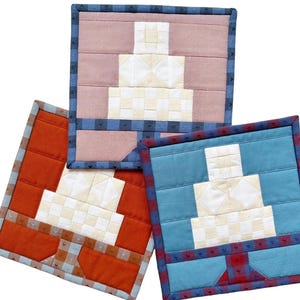 May include: Three square fabric pot holders featuring a pieced quilt design of a tiered cake on a pedestal. The cakes are white and cream, set against backgrounds of dusty rose, burnt orange, and blue, all finished with patterned borders.