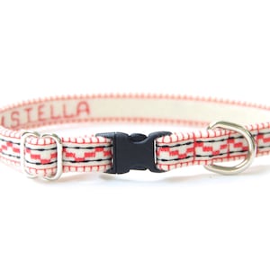 May include: A white and red patterned dog collar with a black plastic buckle and a silver metal D-ring. The collar is personalised with the name "STELLA" in red thread.