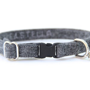 May include: Grey felt cat collar featuring a black plastic buckle, silver-tone hardware, and a small bell. The word "STELLA" is faintly visible on the collar. A simple and stylish pet accessory.