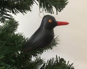 May include: A black bird Christmas ornament with a red beak and yellow eyes hangs on a green Christmas tree. The bird is made of a smooth material and has a simple, elegant design. The ornament is suspended by a metal hook.