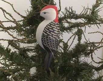 May include: A hand-painted wooden ornament of a red-bellied woodpecker. The bird has a red head, white belly, and black and white patterned back. It hangs from a green string and a gold-colored wire loop, set against a backdrop of a green Christmas tree.