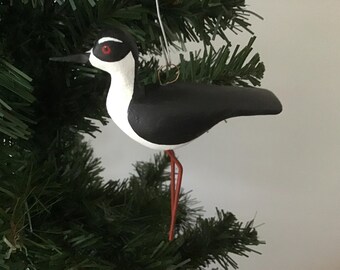 May include: A hand-painted bird ornament with a black body, white chest, and a black head with a white stripe. The bird has red eyes and long, orange legs. It is hanging from a Christmas tree.