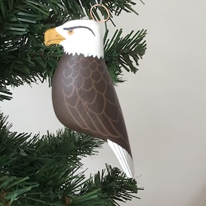 May include: A wooden bald eagle Christmas ornament. The eagle has a white head, a yellow beak, and brown body with white wing tips. The ornament is hanging on a green Christmas tree.