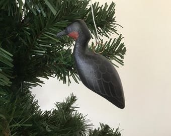 May include: A black bird-shaped Christmas ornament with a red throat hangs from a green Christmas tree. The bird has detailed wing and feather patterns. The ornament is suspended by a gold-colored wire.