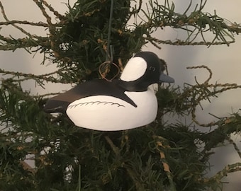May include: A hand-painted wooden ornament of a bufflehead duck. The duck is white with a black head and a black stripe around its neck. The ornament is hanging from a tree branch.