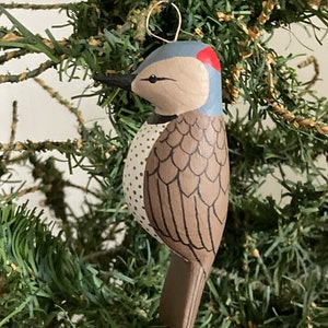 May include: A hand-painted wooden bird ornament with brown, white, and blue paint. The bird has a red crown and a brown tail. The ornament is hanging from a green artificial Christmas tree.