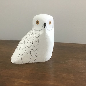 May include: White wooden owl figurine with black eyes and a black beak. The owl has a detailed design of feathers on its body.
