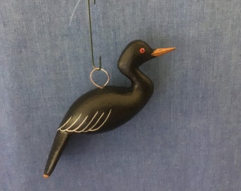 May include: A hand-carved wooden bird ornament. The bird is painted black with a light orange beak and red eyes. White lines detail the wings. A small gold-colored wire loop is attached for hanging. The ornament hangs from a green wire.