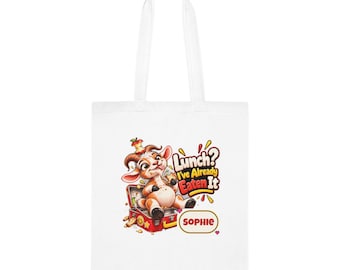 Può includere: Una borsa tote bianca con una capra dei cartoni animati seduta in un portapranzo rosso con un torsolo di mela sulla testa. Il testo recita "Lunch? I've Already Eaten It" e il nome "SOPHIE" è stampato in un ovale giallo sotto il disegno.