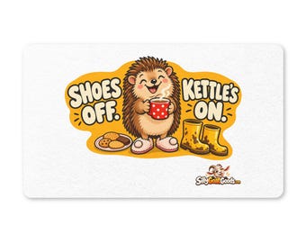 Könnte beinhalten: Eine rechteckige weiße Fußmatte mit einem Cartoon-Igel, der eine rote Tasse mit weißen Punkten hält, flauschige Hausschuhe trägt, neben schlammigen gelben Stiefeln und einem Teller Kekse. Der Text lautet SHOES OFF. KETTLE'S ON. und zeigt das Logo SillyGoatGoods.com.