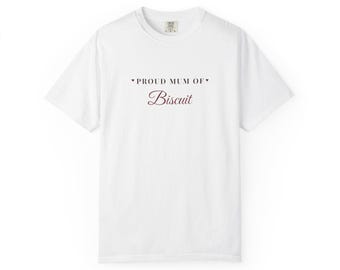 May include: A white short-sleeve t-shirt featuring the text PROUD MUM OF in a dark sans-serif font, flanked by two small dark hearts, with the word Biscuit written below in a dark red cursive script font centered on the chest area.