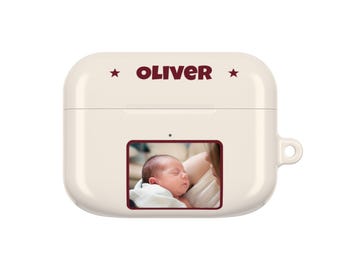 Op de afbeelding: Een crèmekleurig beschermhoesje voor draadloze oordopjes met de naam OLIVER in donkerrode tekst, geflankeerd door twee kleine sterren. Onder de naam bevindt zich een rechthoekige fotoafdruk van een slapende pasgeboren baby in een witte deken.