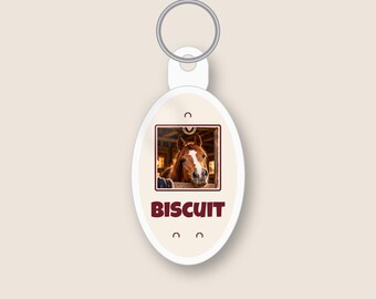 May include: An oval-shaped white keyring featuring a central square photograph of a brown horse with a white blaze on its face, positioned inside a stable. The name BISCUIT is printed in bold dark red capital letters below the photograph.